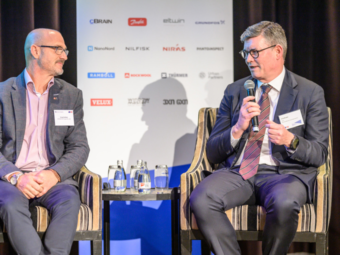 Deux hommes en costume discutent sur scène, avec un micro, des bouteilles d'eau et un panneau publicitaire en arrière-plan.