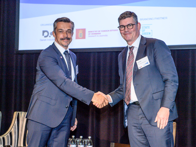 Deux hommes en costume se serrant la main lors d'un événement officiel, avec un écran de projection et des bouteilles d'eau en arrière-plan.
